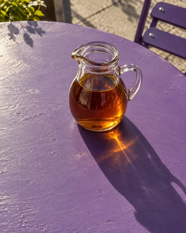 An open clear glass jug of amber syrup sits on a violet-painted outdoor café table, with a long diagonal shadow and a bright amber reflection beneath it.