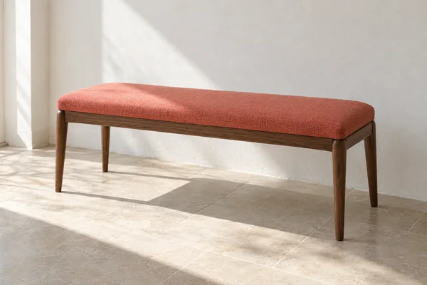 A coral-upholstered wooden bench stands against a pale plaster wall on a light stone-tile floor, lit by strong daylight from the left.