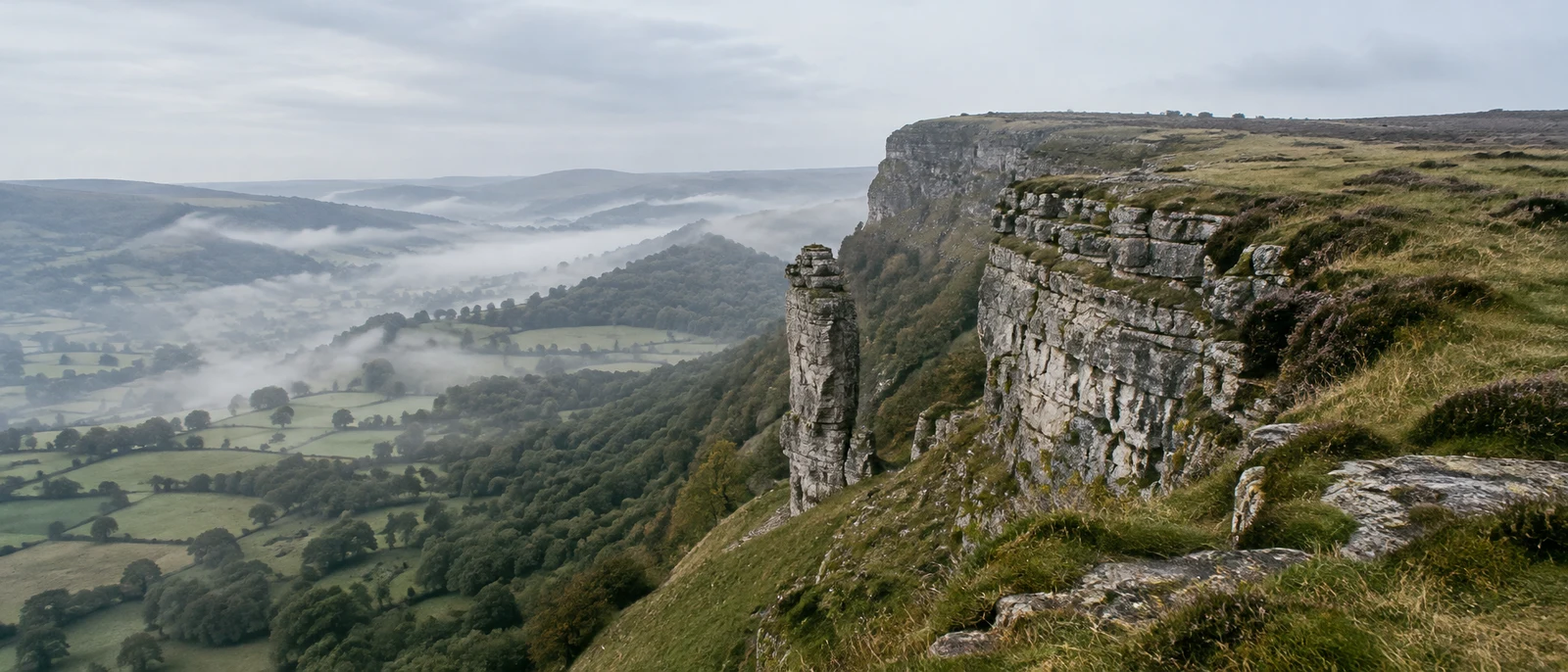 A narrow limestone rock fin stands beside a stepped escarpment above a mist-filled valley of patchwork fields and woodland, with grassy ground in the…