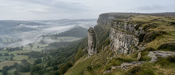 A narrow limestone rock fin stands beside a stepped escarpment above a mist-filled valley of patchwork fields and woodland, with grassy ground in the…