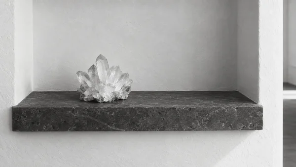 A translucent quartz crystal cluster rests left of center on a dark stone ledge inside a white plaster recess, shown in monochrome with soft light and visible…