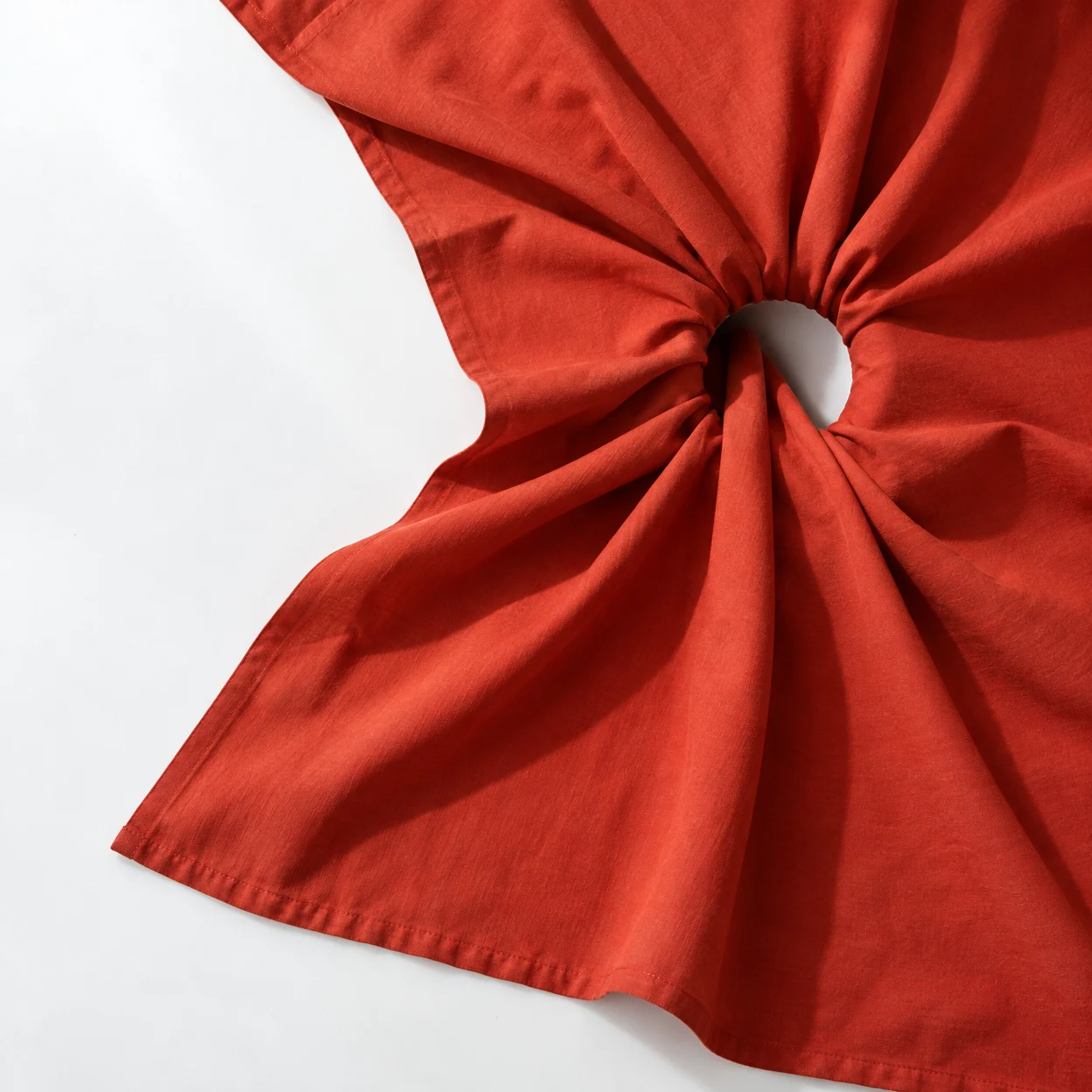 Overhead view of vermilion fabric gathered around a dark circular opening, spreading into irregular folds across a white surface; the curved stitched hem runs…