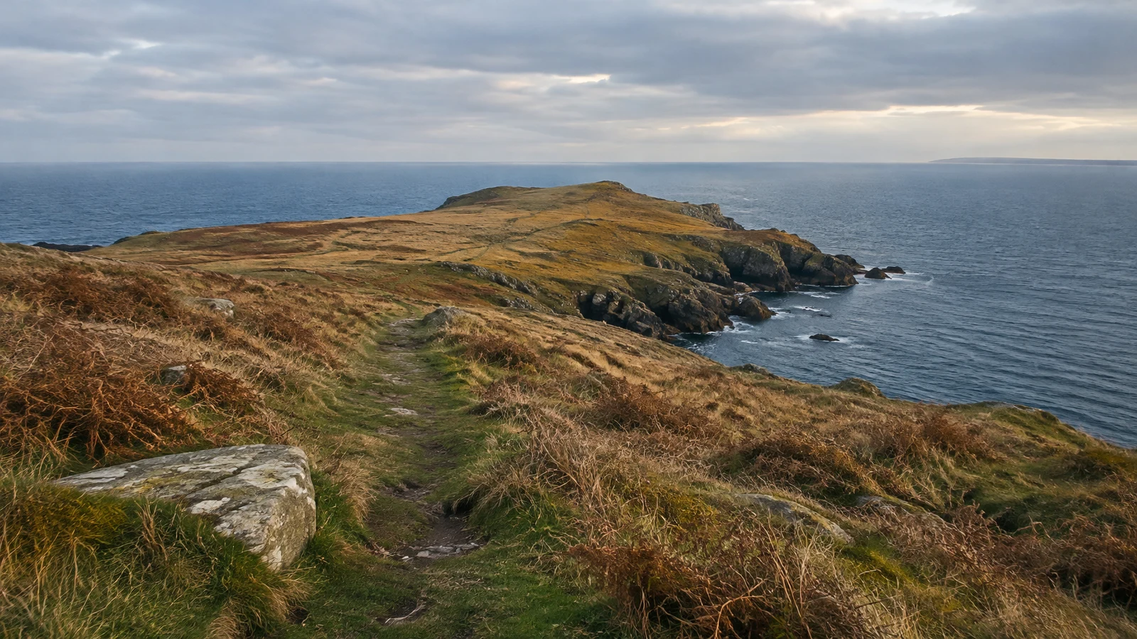 A narrow worn path crosses tawny grass and bracken toward a rocky coastal headland, with a broad weathered stone in the foreground and blue sea beneath a…