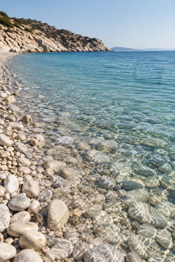 A clear Mediterranean-looking bay with pale rounded pebbles in the foreground, transparent shallows over submerged stones, and a rocky scrub-covered coast…