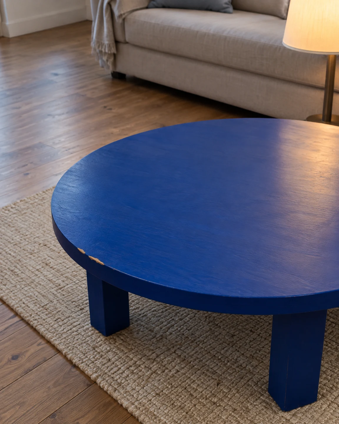 A low cobalt-blue round coffee table with small chips exposing pale wood along its rim sits on an oatmeal rug over timber flooring; a cream-shaded floor lamp…