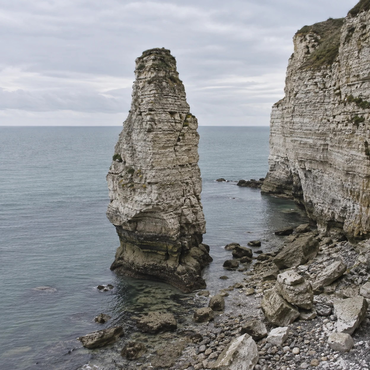 A tall, layered limestone sea stack rises from calm grey-blue water beside a pale stratified cliff, with broken rocks and pebbles along the shore beneath an…
