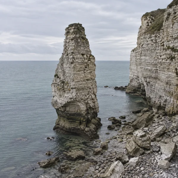 A tall, layered limestone sea stack rises from calm grey-blue water beside a pale stratified cliff, with broken rocks and pebbles along the shore beneath an…