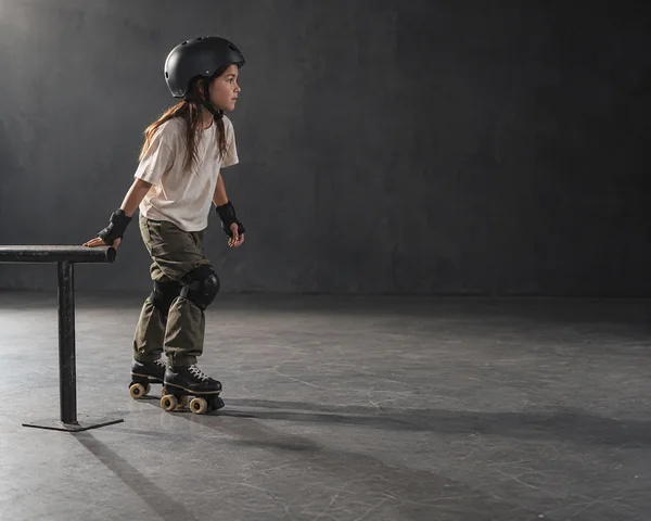 A child wearing a black helmet, wrist guards and knee pads stands on quad skates beside a low metal practice rail in a dark studio, with open grey floor to the…