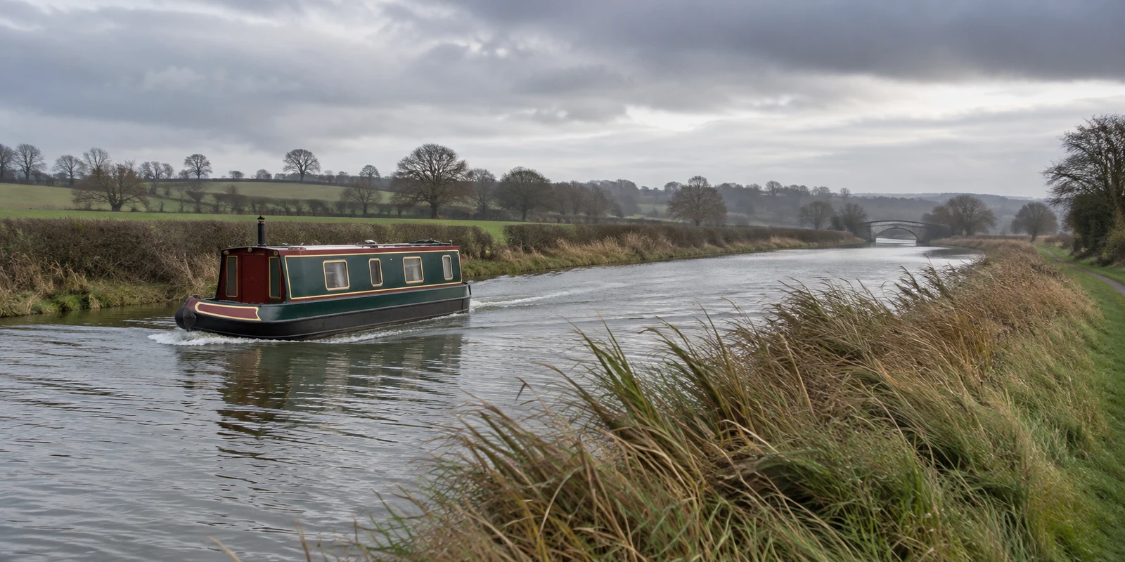 A traditional narrowboat with a muted green hull and faded red trim cruises along a grey-green canal, leaving a small wake.