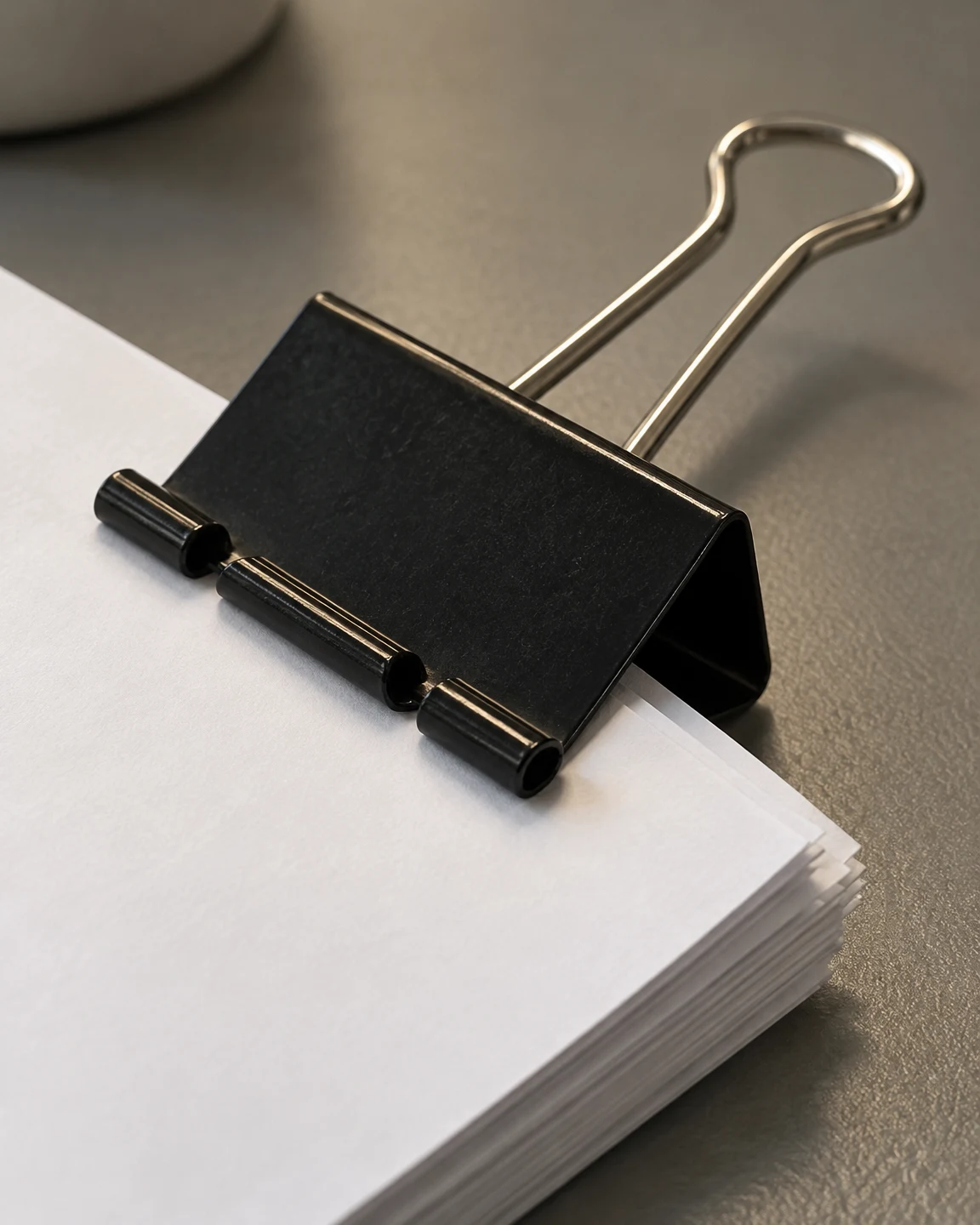 A black binder clip grips a thick diagonal stack of blank white paper on a textured grey desk, with a silver wire handle extending toward the upper right and a…