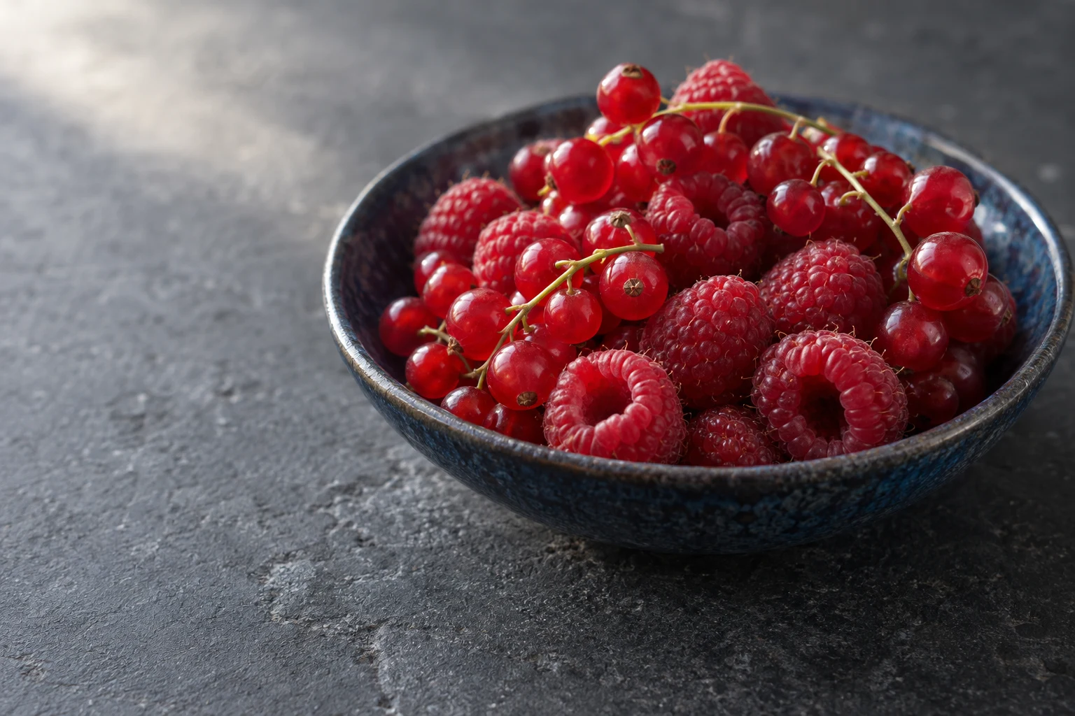 A large indigo ceramic bowl filled with redcurrants on stems and raspberries sits on a charcoal stone surface, with open space to the left.