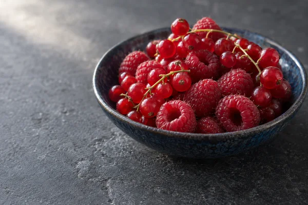 A large indigo ceramic bowl filled with redcurrants on stems and raspberries sits on a charcoal stone surface, with open space to the left.