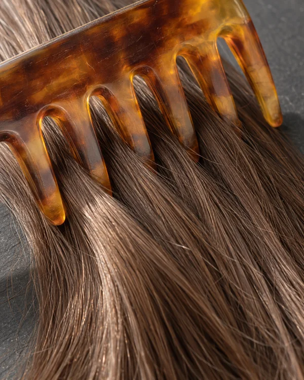 Extreme closeup of a translucent amber comb passing through chestnut-brown hair on a dark graphite surface, with fine strands visible between the rounded teeth.