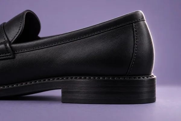 Extreme close-up of a black leather loafer, showing its rear quarter, stitched welt and layered block heel against a pale-violet tabletop and matching…