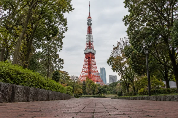 A low view along a brick-paved path in a tree-lined park toward Tokyo Tower beneath a pale overcast sky, with modern buildings visible behind the trees.