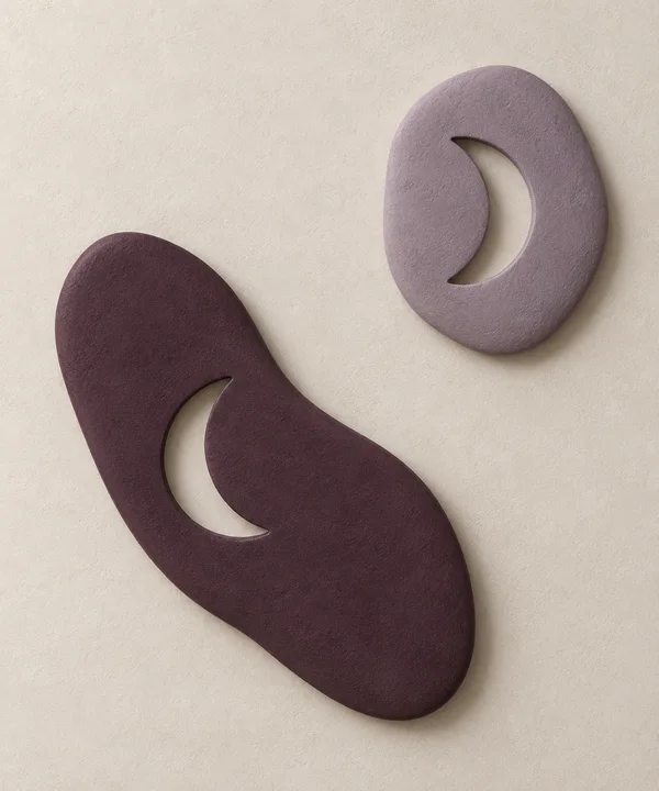 Two matte sculptural reliefs sit diagonally on a pale textured ground: a large dark aubergine form at lower left and a smaller dusty-lilac oval at upper right…
