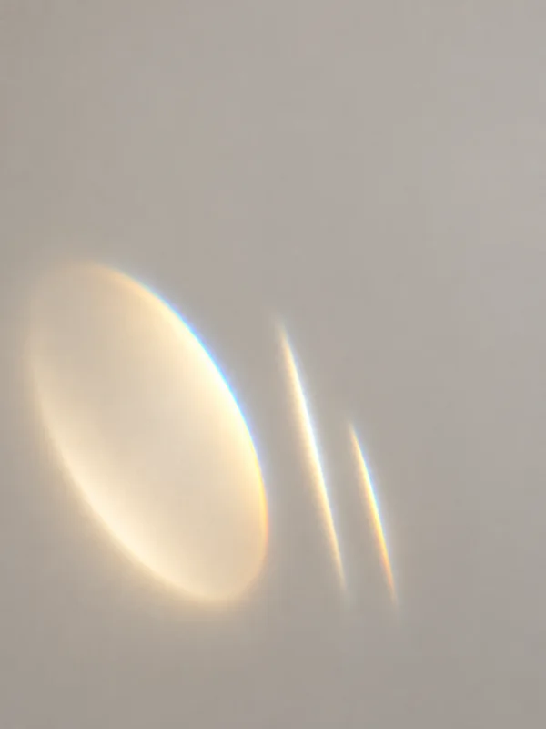 Three luminous forms sit in the lower-left area of a warm grey field: a large tilted pale oval with a cyan-and-amber edge, and two slender, slightly curved…