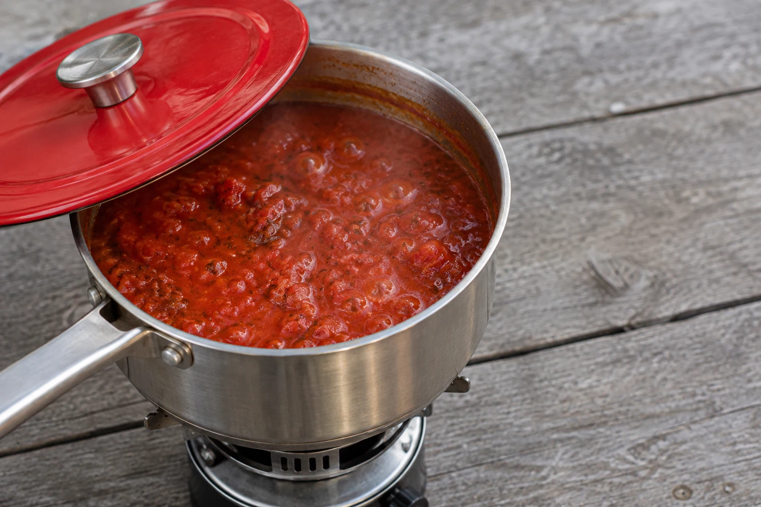 A stainless-steel saucepan of bubbling red tomato sauce sits on a compact burner on a weathered grey wooden table, with a red lid slid diagonally across the…