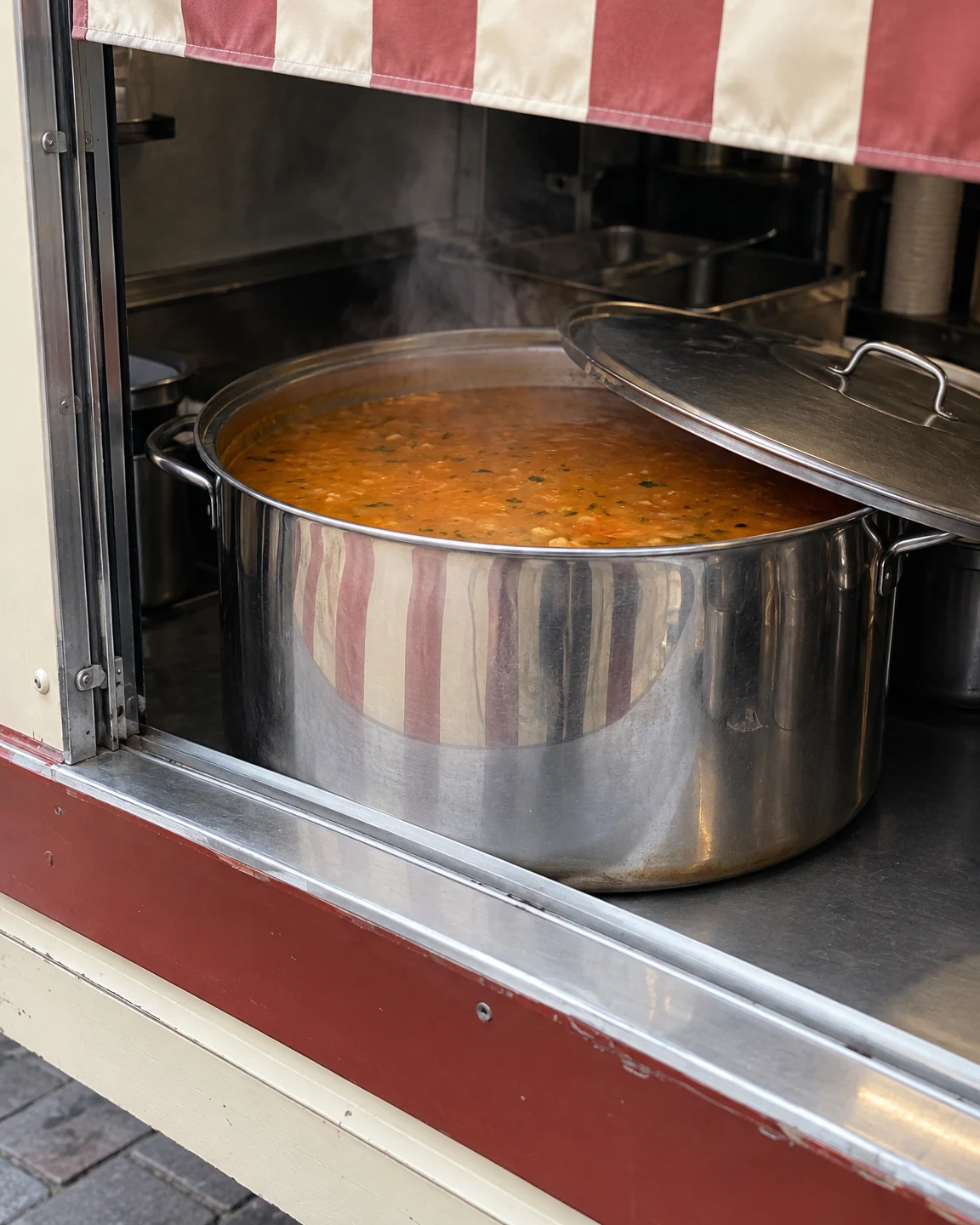 A large stainless-steel pot of orange soup sits in a street-food stall’s serving hatch, with the lid slid partly aside and steam rising.
