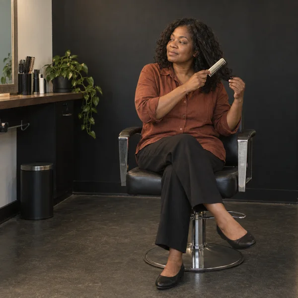 A middle-aged woman with shoulder-length dark curls sits cross-legged in a black hydraulic salon chair, combing a section of her hair with a wide-tooth comb.