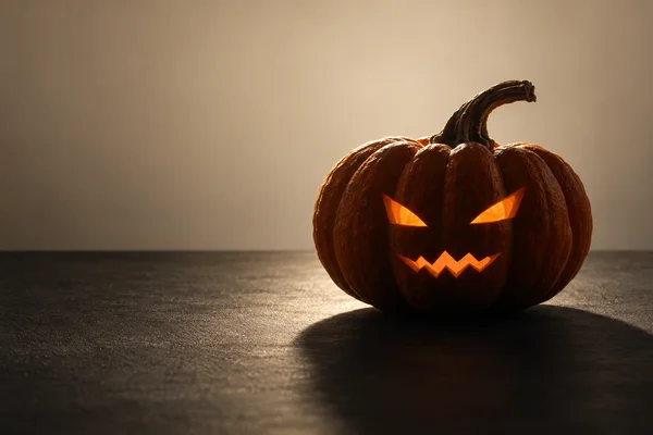 A deeply ribbed orange jack-o'-lantern with glowing angular eyes and a zigzag mouth sits on a dark tabletop against a warm cream background, casting a long…