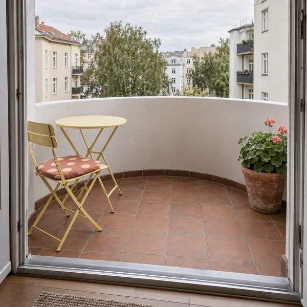 View through an open apartment doorway onto a small balcony with terracotta tiles, a curved white plaster parapet, a yellow folding chair and round table, a…