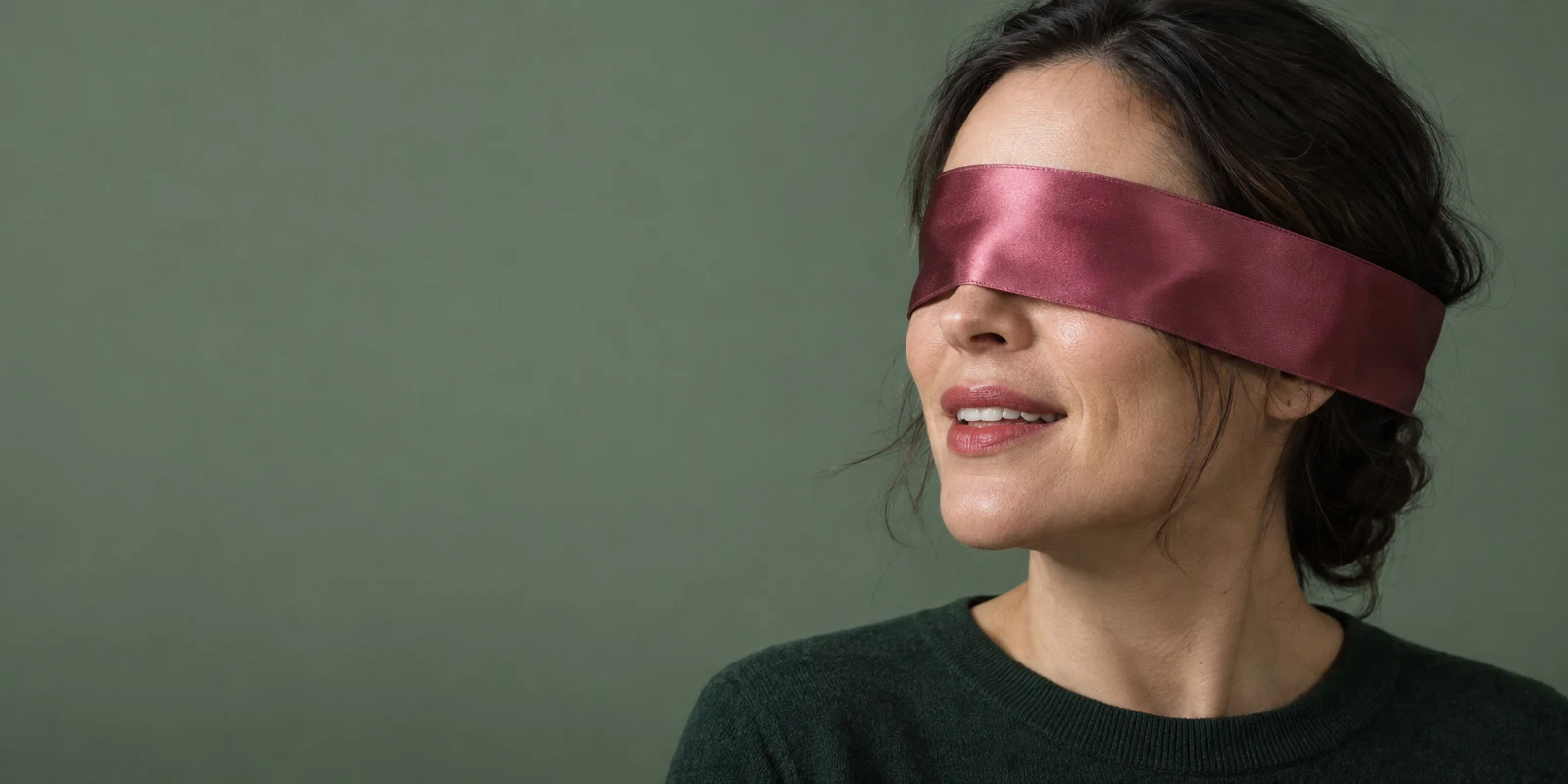 A woman in a dark green crewneck is framed on the right against a muted sage-green background, her eyes covered by a broad dusty-magenta satin ribbon.
