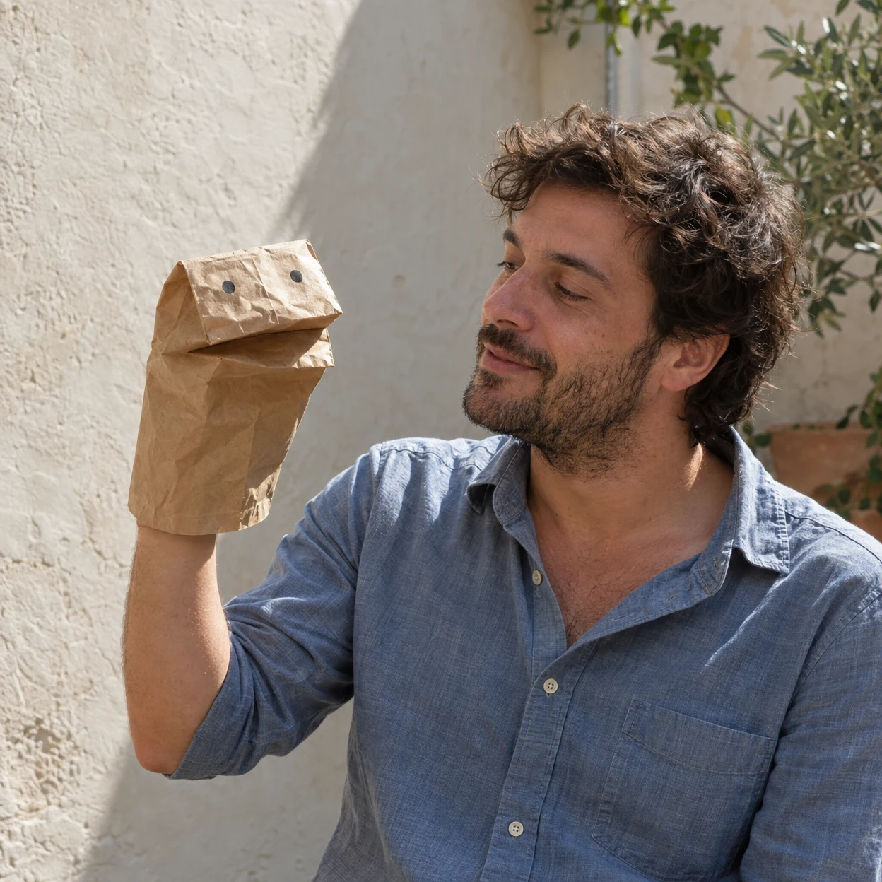 A man in a faded blue shirt holds a creased kraft paper bag puppet with two dark eyes and a folded mouth, looking toward it in a sunlit plaster-walled…