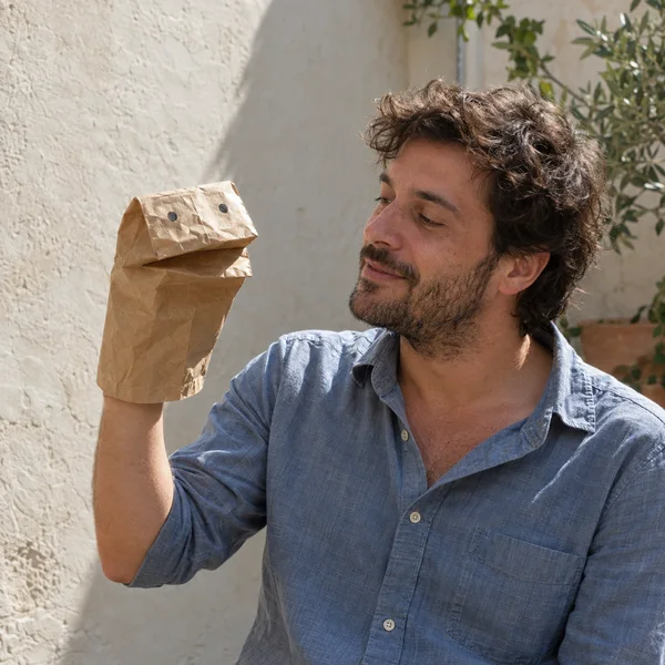 A man in a faded blue shirt holds a creased kraft paper bag puppet with two dark eyes and a folded mouth, looking toward it in a sunlit plaster-walled…