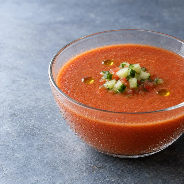 A large clear glass bowl of orange-red tomato gazpacho sits on a blue-gray stone surface, topped with diced cucumber and three olive oil droplets.