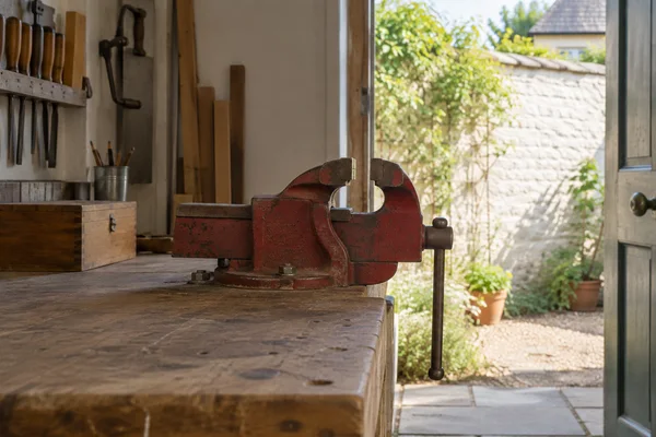 A worn red bench vise sits on a wooden workbench inside a small workshop, with hand tools on the wall and an open doorway onto a sunlit garden.