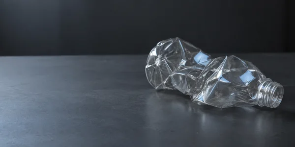 A clear, uncapped plastic bottle with a label-free, heavily crushed body lies on its side across the right half of a charcoal tabletop, lit by cool rectangular…