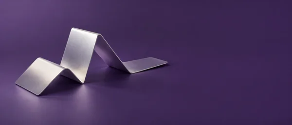 A broad satin-silver metal strip folds into a sculptural zigzag on a muted violet surface, positioned left of center with open space to the right.