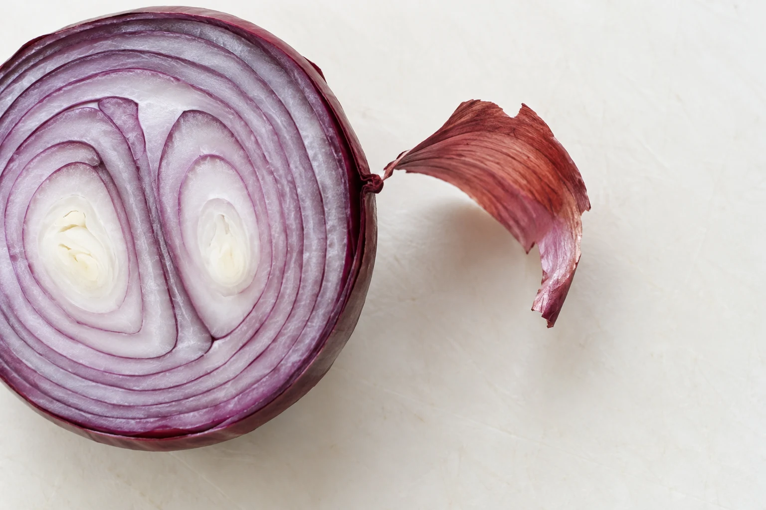 Overhead close-up of a halved red onion on an ivory board, its purple-and-white cut rings filling the left side and a strip of reddish-purple skin curling into…