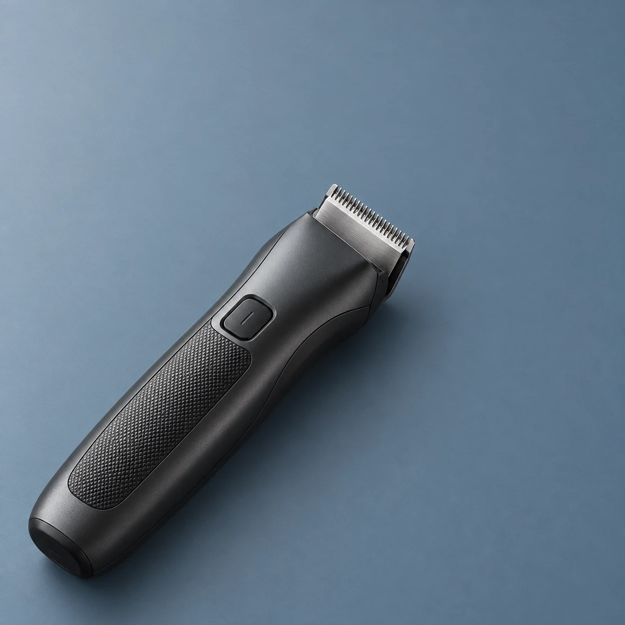 A matte graphite cordless hair trimmer rests diagonally on a muted blue-grey surface, with its stainless-steel cutting head pointing upper right and a textured…