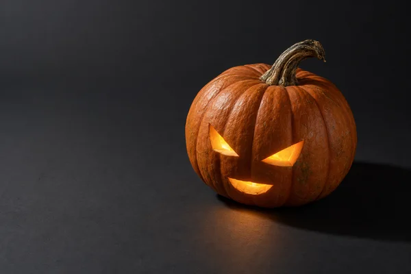 A glowing burnt-orange jack-o’-lantern with angular eyes and a curved, toothless mouth sits on a charcoal tabletop against a dark seamless background, with…