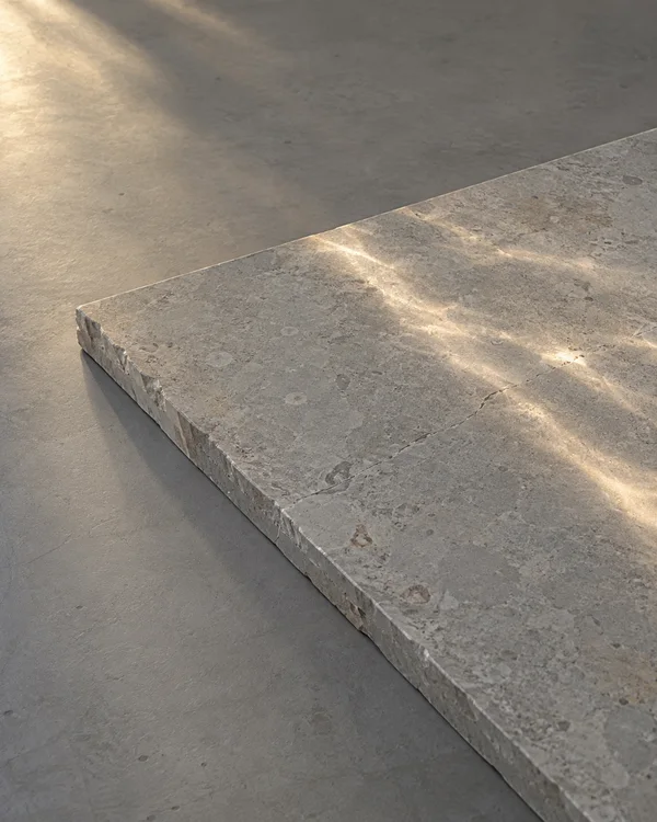 A thick grey limestone slab lies diagonally across a matte grey tabletop, with a rough cut edge and warm bands of reflected light crossing its surface.