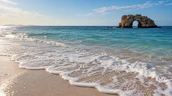 A low view across a pale sandy beach as a scalloped wave of white foam spreads over wet sand, with blue-green water, open sky and a weathered limestone sea…