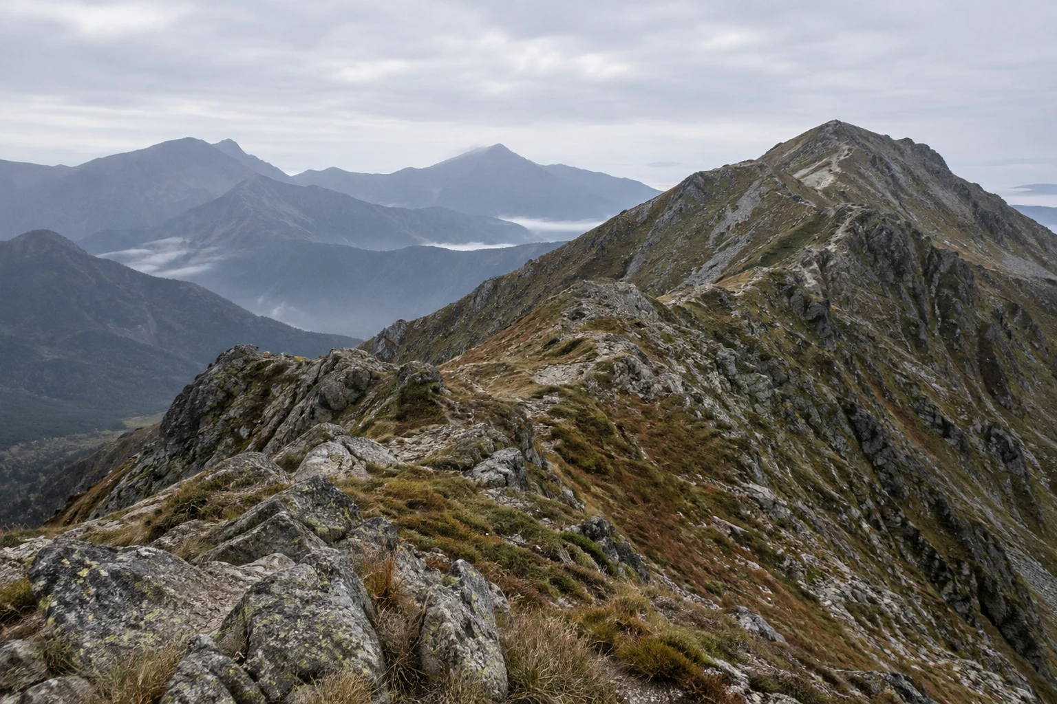A rugged, lichen-spotted rocky ridge with muted alpine grasses climbs toward a summit on the right, with layered blue-grey mountains and mist-filled valleys…