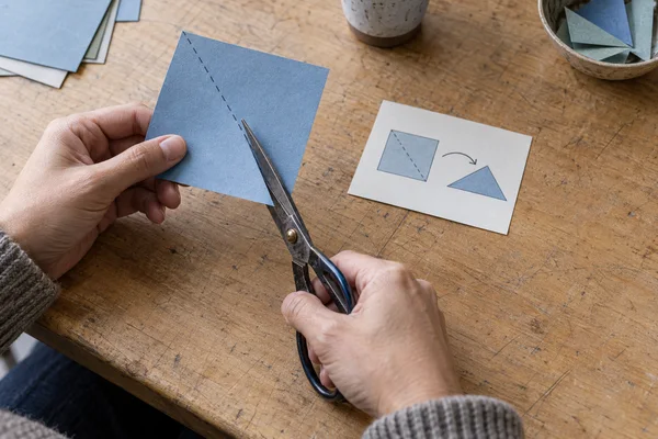 Two hands hold a blue paper square and worn scissors over a scratched wooden table, with the blades positioned along a dashed diagonal guide.
