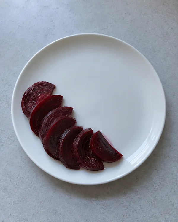 Seven dark burgundy beetroot slices overlap in a fan along the lower-left of a white plate on a pale gray counter, viewed from above.
