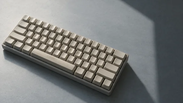 A blank cream mechanical keyboard rests diagonally on a pale blue-grey tabletop, with shiny patches on several keys and a broad shadow across the open space to…