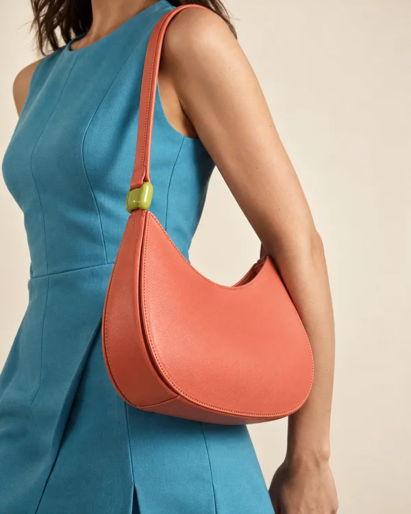 A cropped fashion model in a cyan sleeveless tailored dress wears a coral crescent shoulder bag with visible edge stitching and a lime-colored clasp against a…
