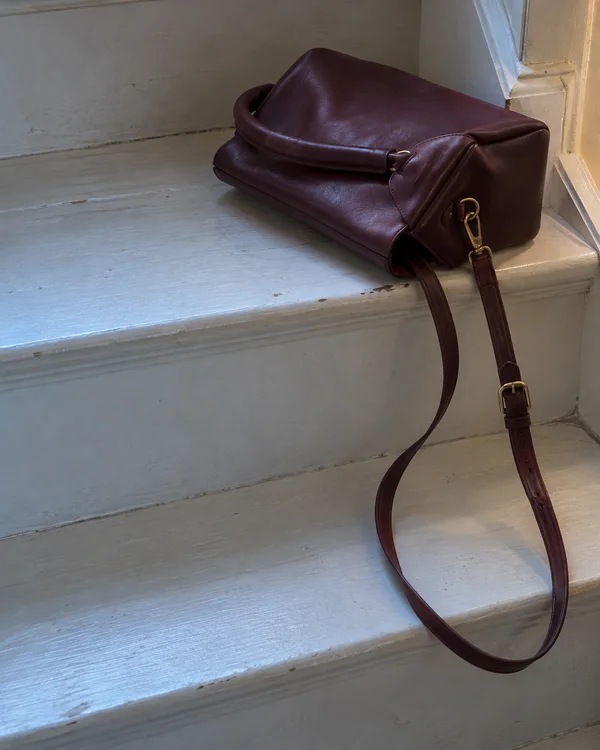 A burgundy leather handbag rests on a worn pale-painted stair tread, with its short handle folded over the bag and a long brass-buckled strap looping down…