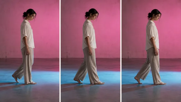 A barefoot woman in loose light trousers and an ivory shirt walks slowly in profile across a studio floor, shown in three side-by-side panels against a pink…