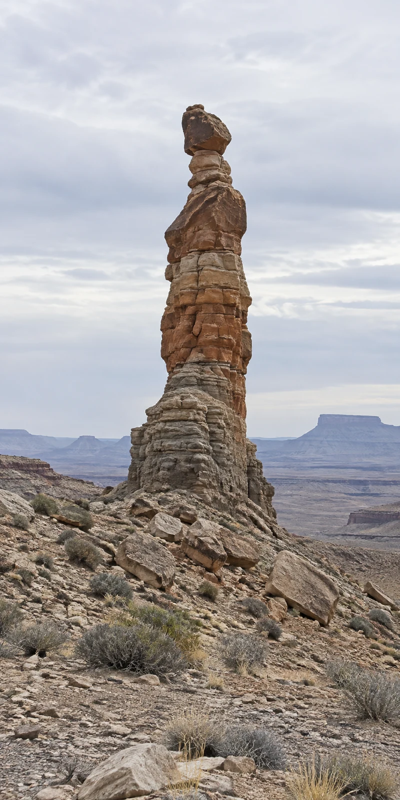 A tall, layered sandstone pinnacle with a dark capstone rises from a rocky desert slope, with sparse shrubs, distant flat-topped mesas and a broad cloudy sky…