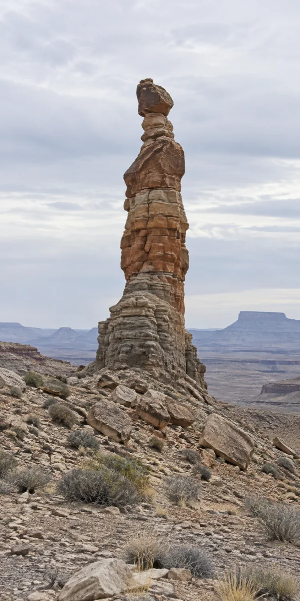 A tall, layered sandstone pinnacle with a dark capstone rises from a rocky desert slope, with sparse shrubs, distant flat-topped mesas and a broad cloudy sky…
