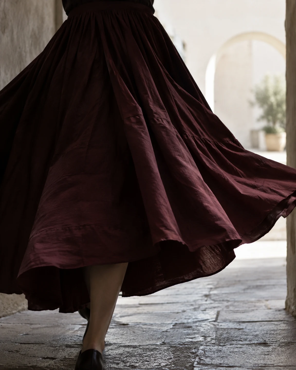 A dark burgundy skirt swings outward around a cropped wearer walking through a stone passage, with one lower leg and dark shoe visible beneath the hem and a…