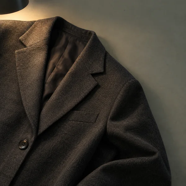 An overhead close view of a dark charcoal jacket laid diagonally on a grey-green tabletop, with its notched lapel and one button prominent.