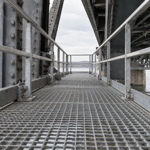 A galvanized square-grid inspection walkway runs beneath a steel bridge toward a pale overcast opening, with metal safety rails and heavy riveted beams framing…